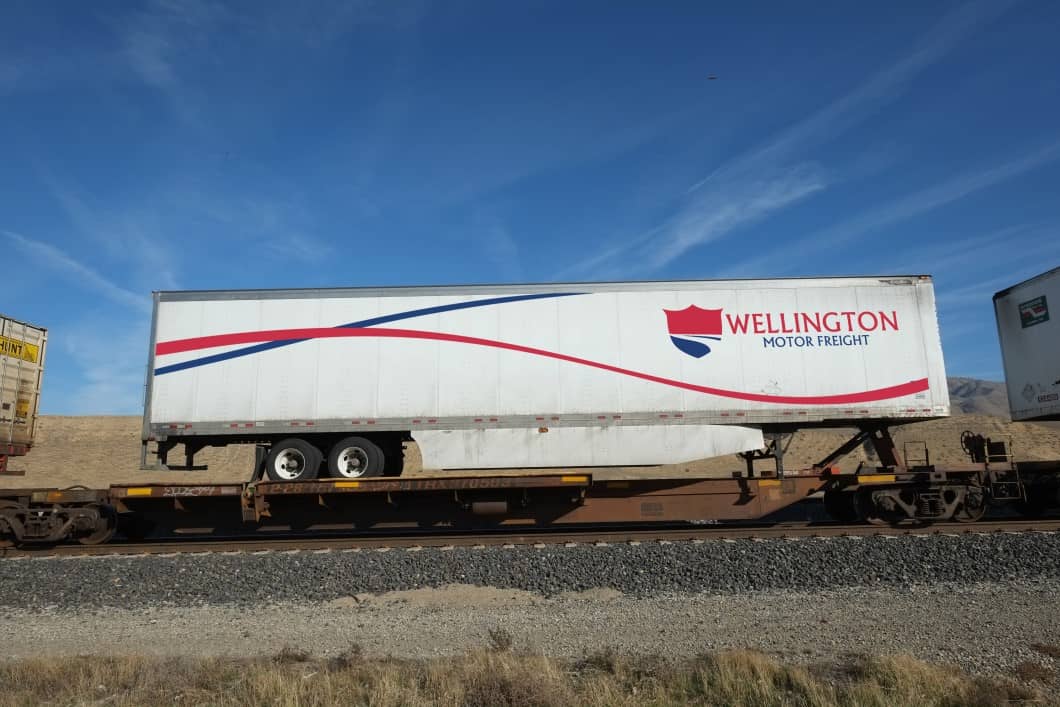 A Wellington Motor Freight 53' trailer ontop of a flat car (TOFC) moving on the railroad.